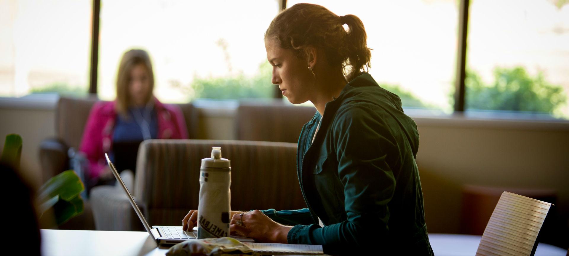 A student studying at a table with a water bottle and laptop as another student sits in the background in front of a window.